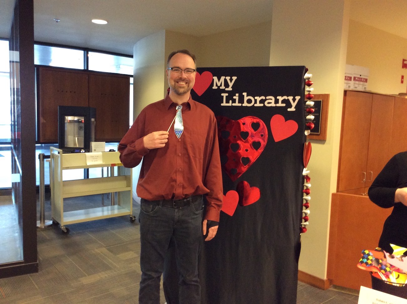 Student success librarian Brad Doerkerson stands in front of a black backdrop covered in red hearts and Love My Library across the top of it. He is holding up a prop necktie on a stick and smiling broadly.
